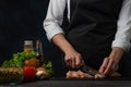 The professional chef in black apron cuts with knife chicken fillet on chopping board, dark blue background. Close-up. Backstage Royalty Free Stock Photo