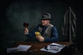 Professional detective in fedora hat sitting at table, drinking red wine and eating fries over dark green vintage Royalty Free Stock Photo