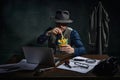Professional detective in fedora hat sitting at table, working on laptop and eating fries over dark green vintage Royalty Free Stock Photo