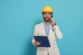 Professional engineer in hard hat with clipboard talking on phone against light blue background Royalty Free Stock Photo