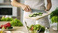 Professional female cook adding fresh cucumber slices in glass bowl with salad Royalty Free Stock Photo