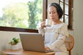 Focused aged Asian businesswoman working from home, sipping coffee while using laptop Royalty Free Stock Photo