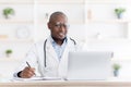 Professional help concept. Friendly black male doctor in white unifrom sitting at table with laptop at his office Royalty Free Stock Photo