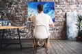 Professional painter sits on chair in front of easel with canvas, hold paintbrush in hand and drawing oil painting. Working proces Royalty Free Stock Photo