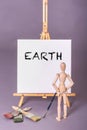 Wooden manikin doll standing in front of canvas with word Earth painted in black