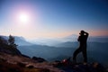 Professional photographer takes photos with mirror camera on peak of rock. Dreamy fogy landscape, spring orange pink misty sunrise Royalty Free Stock Photo