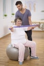 Professional physiotherapist stabilizing senior woman sitting on exercising ball