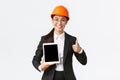 Professional smiling asian female construction manager, factory engineer in business suit and safety helmet showing