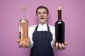 Professional waiter in uniform holds two bottles of red and white wine, looks at them and rejoices, smiles against a pink Royalty Free Stock Photo