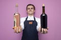Professional waiter in uniform holds two bottles of red and white wine and smiles on a pink background, the sommelier gives and Royalty Free Stock Photo