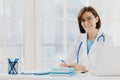 Professional woman doctor writes down notes, poses at desktop in office with laptop, wears white coat, spectacles and Royalty Free Stock Photo