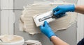 Professional hands in blue gloves spreading tile adhesive with a notched trowel on a grey wall surface. Royalty Free Stock Photo