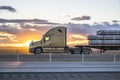 Profile of big rig beige semi truck transporting cargo on flat bed semi trailer driving on the one way highway road at sunset time