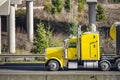 Profile of bright yellow classic bonnet big rig semi truck long haul tractor with black covered semi trailer driving on the