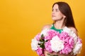 Profile of brunette young lady wearing green clothing, embracing huge bouquet of peonies, female looking aside, standing against