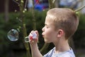Profile of cute handsome small blond child boy with funny serious expression blowing colorful transparent soap bubbles outdoors o Royalty Free Stock Photo