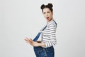 Profile of excited brunette girl with two hairbuns with opened mouth, has cheerful expression, wears denim overalls