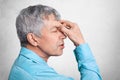 Profile of exhausted grey haired male in formal shirt, keepd hands on nose, poses against white concrete background, tries to conc Royalty Free Stock Photo