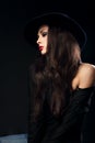 Profile of expressive female model posing in black shirt and elegant hat with red bright lipstick on dark shadow background.