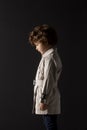 Profile of fashionable adorable curly little boy, wear in trench, poses with his head down, over black background