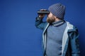 Bearded man in a knitted hat and a raincoat looks through binoculars, standing on an isolated blue background. Copy Royalty Free Stock Photo