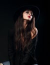 Profile of glamour female model posing in black shirt and elegant hat with red lipstick on dark shadow background and looking up.