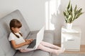 Profile indoor shot of little cute dark haired female child wearing white casual t shirt using laptop while sitting on couch,