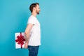 Profile portrait of cheerful young man arms hold giftbox behind back look empty space isolated on blue color background
