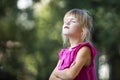 Profile portrait of cute adorable blond child girl in sleeveless pink dress outdoors with raised head and closed eyes on blurred g Royalty Free Stock Photo