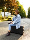 Profile portrait pensive girl sits on her suitcase on middle road, waiting a transportation, in sunset background. Royalty Free Stock Photo