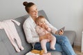 Profile portrait of positive smiling woman wearing white shirt and jeans sitting on sofa with baby daughter, holding cell phone Royalty Free Stock Photo