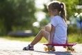 Profile portrait of pretty small long-haired blond girl in white clothing sitting on skateboard on bright summer day on blurred