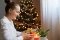 Profile portrait of smiling beautiful woman wearing white shirt sitting on table near christmas tree and holding present box in Royalty Free Stock Photo