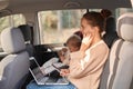 Profile portrait of woman wearing beige jumper sitting with her baby daughter in safety chair on backseat of car, holding mobile