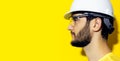 Profile portrait of young bearded man wearing construction safety helmet and glasses on yellow background. Royalty Free Stock Photo
