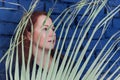 Profile portrait young red-haired woman behind large branch of palm tree on blue brick wall background Royalty Free Stock Photo