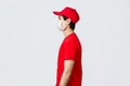 Profile shot of asian delivery guy in red cap and t-shirt, wearing protective medical mask, looking left, waiting at Royalty Free Stock Photo