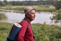 Profile shot of eldery man with backpack and blue rug, wearing red casual sweater and cap, enjoing beautifulnature near river and