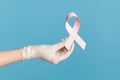Profile side view closeup of human hand in white surgical gloves shwoing and holding pink ribbon, symbol of breast cancer