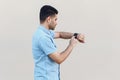 Profile side view portrait of attentive handsome young bearded man in blue shirt standing and using his smart watch, touching at Royalty Free Stock Photo