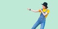 Profile side view portrait of pretty young hipster girl in blue denim overalls, yellow shirt and black hat standing with fist or Royalty Free Stock Photo