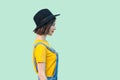 Profile side view portrait of pretty young hipster girl in blue denim overalls, yellow shirt and black hat standing, smiling and Royalty Free Stock Photo