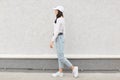 Profile of stylish girl wearing jeans, shirt and baseball cap, looking away, posing outdoor against white wall, woman with dark Royalty Free Stock Photo