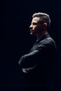 Profile view of confident man in black suit with arms crossed on dark background