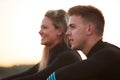 Profile View Of Couple Wearing Wetsuits On Surfing Staycation Sitting On Beach Looking Out To Sea