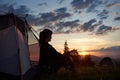 Profile of young woman sitting in tent in grass and fieldflowers on top mountain with view of rising sun