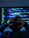 A programmer sits in a dark room, hands clasped over his head in frustration. The glow of the code on the screens