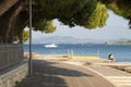 Promenade with pine trees by the sea and people and boats in the distance