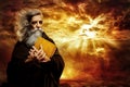 Prophet with Bible. Old Monk with Golden Book praying over Epic Landscape Background. Senior Bearded Man Worship in Black Cloak Royalty Free Stock Photo