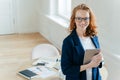 Prosperous female leader of working team holds digital tablet device, develops business ideas, has toothy smile, red hair, wears